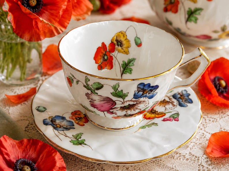 K&ouml;nnte beinhalten: Wei&szlig;es Tee-Set mit Blumendesign aus roten, gelben und blauen Blumen. Die Tasse und Untertasse haben einen goldenen Rand. Rote Mohnblumen sind im Hintergrund.