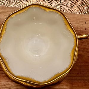 May include: A white teacup with a gold rim and a scalloped edge. The cup is empty and sitting on a wooden surface.