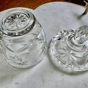 May include: A clear glass butter dish with a faceted starburst pattern on the base and lid. The lid features a rounded knob handle, and the base has a circular rim. Both pieces are made of transparent glass with intricate geometric cut designs.