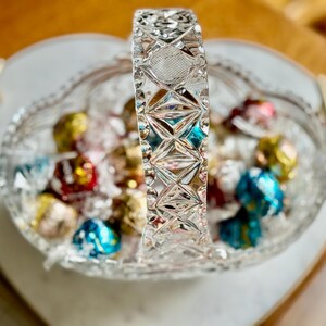 May include: A clear, heart-shaped crystal candy dish with a decorative handle, filled with colorful wrapped candies. The handle features a diamond pattern, adding to the dish's elegant design. The candies are in various colors.