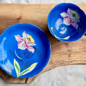 Puede incluir: Un juego de taza y platillo de t&eacute; azul y blanco con un dise&ntilde;o floral. La taza y el platillo tienen un borde festoneado y est&aacute;n decorados con una flor rosa con detalles amarillos y ribetes dorados.
