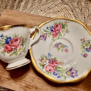 May include: A white teacup and saucer set with a floral pattern in shades of pink, purple, and yellow. The set has a gold trim around the edges.