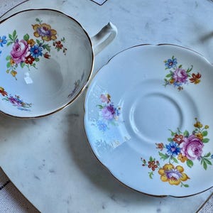 Puede incluir: Un juego de taza y platillo blancos con un dise&ntilde;o floral. La taza y el platillo tienen un borde dorado y est&aacute;n decorados con flores rosas, azules y amarillas. El juego est&aacute; colocado sobre una bandeja de m&aacute;rmol blanco.