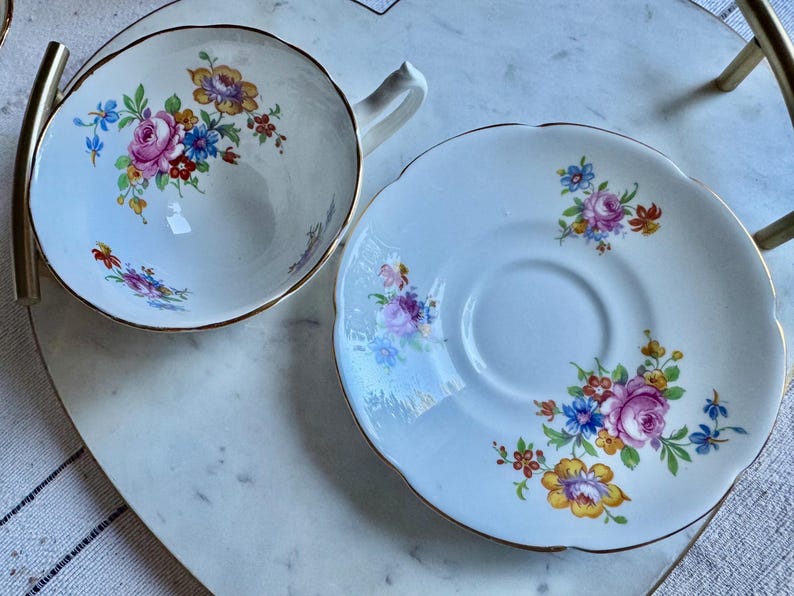 Puede incluir: Un juego de taza y platillo blancos con un dise&ntilde;o floral. La taza y el platillo tienen un borde dorado y est&aacute;n decorados con flores rosas, azules y amarillas. El juego est&aacute; colocado sobre una bandeja de m&aacute;rmol blanco.