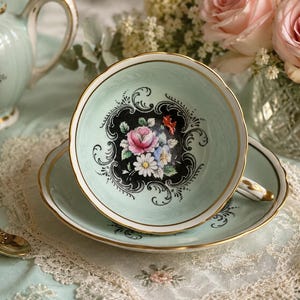 May include: A pale mint green porcelain teacup and saucer set featuring a central black medallion with a floral bouquet of pink roses and white daisies, accented by intricate black scrollwork and a delicate gold rim on the edges.
