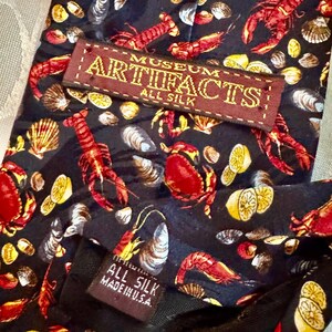 Puede incluir: Una corbata de seda negra con un patr&oacute;n repetitivo de langostas rojas, cangrejos, camarones, varias conchas marinas y rodajas de lim&oacute;n amarillo. Una etiqueta marr&oacute;n dice MUSEUM ARTIFACTS ALL SILK y una etiqueta negra peque&ntilde;a dice ALL SILK MADE IN U.S.A.