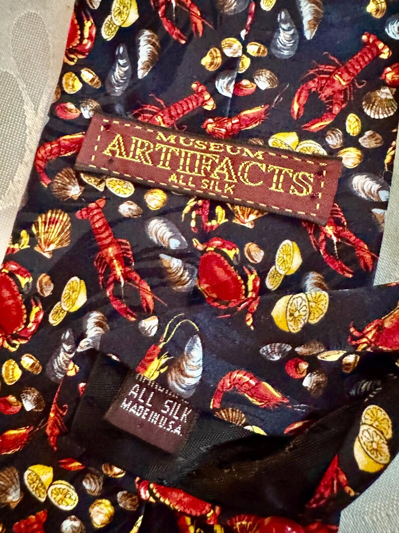 Puede incluir: Una corbata de seda negra con un patr&oacute;n repetitivo de langostas rojas, cangrejos, camarones, varias conchas marinas y rodajas de lim&oacute;n amarillo. Una etiqueta marr&oacute;n dice MUSEUM ARTIFACTS ALL SILK y una etiqueta negra peque&ntilde;a dice ALL SILK MADE IN U.S.A.