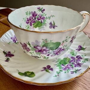 K&ouml;nnte beinhalten: Wei&szlig;es Tee-Service mit lila und gr&uuml;nen Veilchenbl&uuml;ten. Die R&auml;nder sind goldfarben. Die Untertasse hat einen gewellten Rand. Die Tasse hat einen goldenen Henkel.