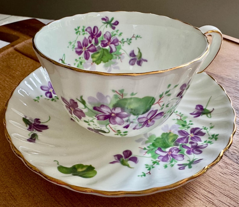 K&ouml;nnte beinhalten: Wei&szlig;es Tee-Service mit lila und gr&uuml;nen Veilchenbl&uuml;ten. Die R&auml;nder sind goldfarben. Die Untertasse hat einen gewellten Rand. Die Tasse hat einen goldenen Henkel.