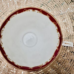 May include: Overhead view of a white teacup with a burgundy rim and gold filigree detailing. The cup has a small handle and sits on a gold-colored placemat. The cup's interior is white with a small, central circle.