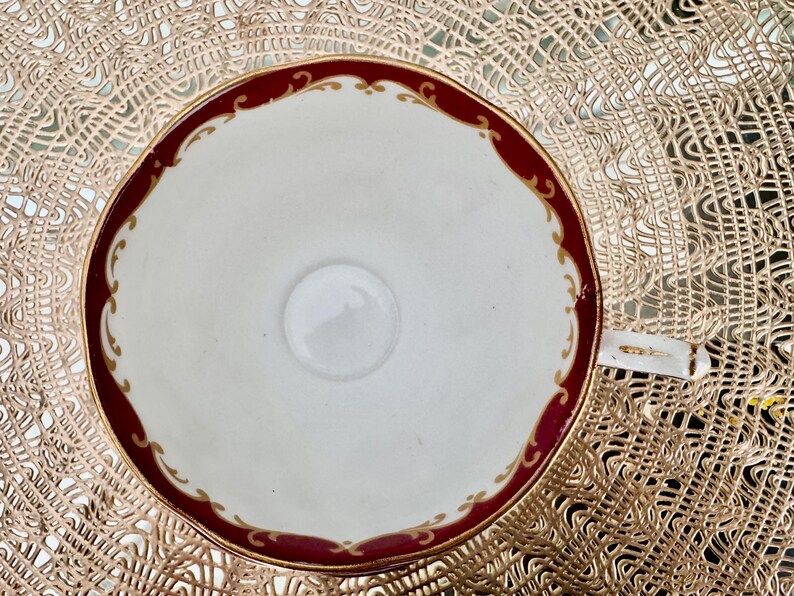 May include: Overhead view of a white teacup with a burgundy rim and gold filigree detailing. The cup has a small handle and sits on a gold-colored placemat. The cup's interior is white with a small, central circle.
