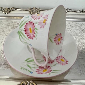 May include: White teacup and saucer set with pink daisy floral design. The teacup has a curved handle and a pink rim. The saucer also has a pink rim and daisy pattern. The set is on a silver tray.