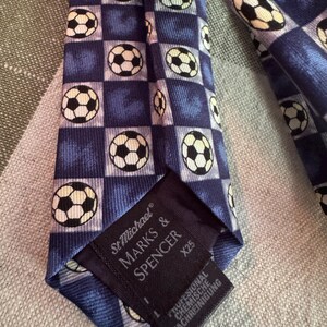 May include: A blue necktie featuring a repeating checkered pattern with white and black soccer ball graphics. The back of the tie includes a black label with the text St Michael MARKS & SPENCER X25 and additional German care instructions.