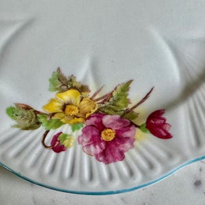 May include: White ceramic plate with a scalloped edge and a light blue trim. The plate is decorated with a floral design featuring yellow and magenta flowers with green leaves. A classic, vintage design.