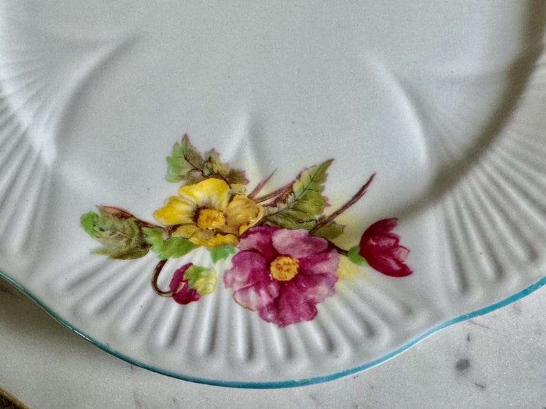 May include: White ceramic plate with a scalloped edge and a light blue trim. The plate is decorated with a floral design featuring yellow and magenta flowers with green leaves. A classic, vintage design.