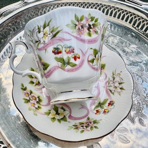 May include: A white teacup and saucer set with floral and ribbon designs. The cup and saucer feature pink ribbons with the names of Canadian provinces. The set is placed on a silver tray.