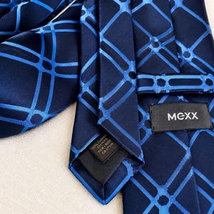 May include: A navy blue tie with a geometric pattern in a lighter shade of blue. The tie is made of a silk blend and features the brand name "MEXX" on a small label.