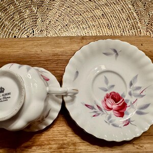 May include: A white teacup and saucer set with a floral design. The cup features a pink rose with gray leaves and the words "Royal Albert Bone China England" on the bottom. The saucer has a similar floral design with a pink rose and gray leaves.
