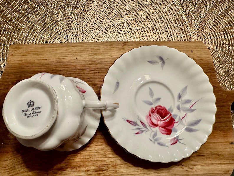May include: A white teacup and saucer set with a floral design. The cup features a pink rose with gray leaves and the words "Royal Albert Bone China England" on the bottom. The saucer has a similar floral design with a pink rose and gray leaves.