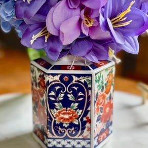 Può includere: Un vaso ottagonale in ceramica con un motivo floreale, con fiori viola e blu. Il vaso ha una base bianca, un bordo blu e rosso a motivi e un disegno centrale di fiori rossi e dettagli blu. Circa 20 cm di altezza.