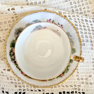 May include: A white porcelain teacup and saucer set featuring a delicate floral pattern with pink roses and green leaves. The edges of both the cup and the saucer are trimmed with a thin gold metallic band. The cup has a small gold-toned handle.