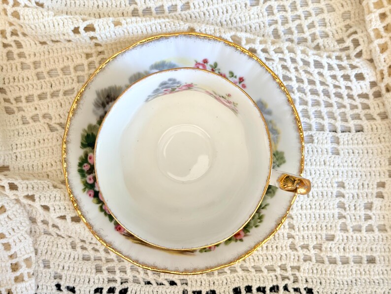 May include: A white porcelain teacup and saucer set featuring a delicate floral pattern with pink roses and green leaves. The edges of both the cup and the saucer are trimmed with a thin gold metallic band. The cup has a small gold-toned handle.