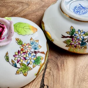 May include: Two porcelain lidded boxes with floral designs. The lid of one box features a three-dimensional pink rose with green leaves. The bottom of the other box has the text "Herend Hungary" in gold lettering.