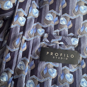 Puede incluir: Una corbata con fondo azul marino oscuro, rayas verticales y un patr&oacute;n repetitivo de motivos florales en azul claro y gris. En la parte posterior hay una etiqueta de tela negra cosida con el texto en blanco PROFILO STUDIO.
