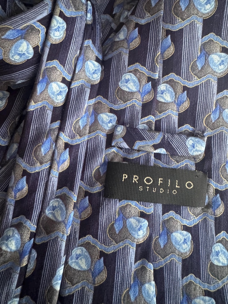 Puede incluir: Una corbata con fondo azul marino oscuro, rayas verticales y un patr&oacute;n repetitivo de motivos florales en azul claro y gris. En la parte posterior hay una etiqueta de tela negra cosida con el texto en blanco PROFILO STUDIO.