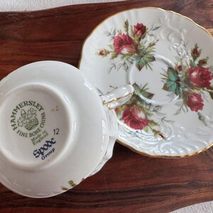 May include: A white teacup and saucer set with a floral design. The saucer has a gold rim and the teacup has a floral design and the text "Hammersley Fine Bone China Made in England Spode Group 12" on the bottom.