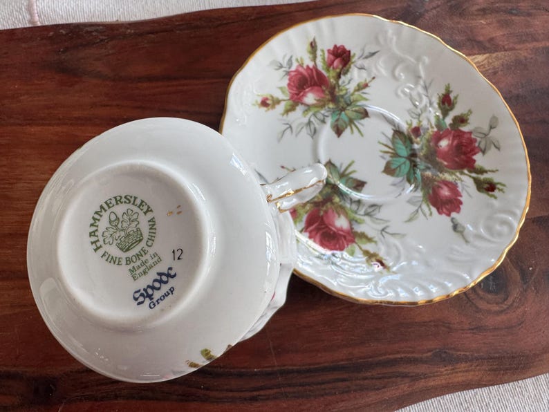 May include: A white teacup and saucer set with a floral design. The saucer has a gold rim and the teacup has a floral design and the text "Hammersley Fine Bone China Made in England Spode Group 12" on the bottom.