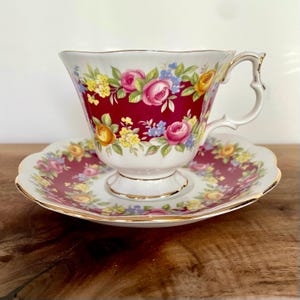 May include: A vintage teacup and saucer set. The teacup features a deep red background with a floral pattern of pink, yellow, and blue roses. The saucer mirrors the design, with gold trim on both pieces. The handle of the teacup has a unique, decorative shape.