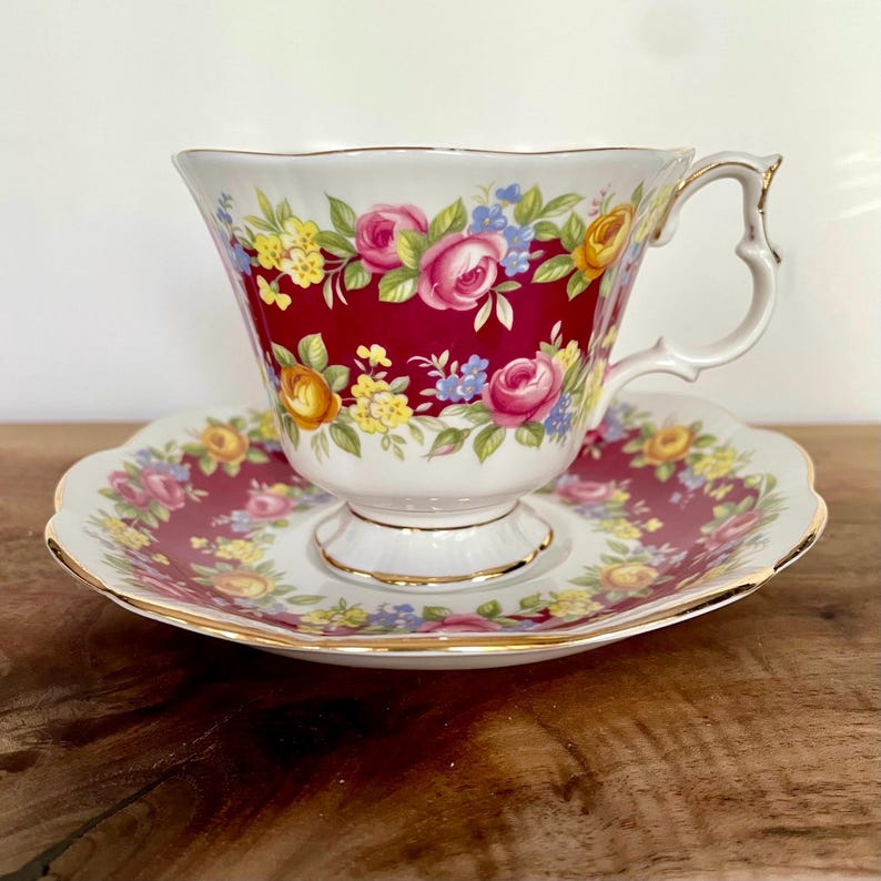 May include: A vintage teacup and saucer set. The teacup features a deep red background with a floral pattern of pink, yellow, and blue roses. The saucer mirrors the design, with gold trim on both pieces. The handle of the teacup has a unique, decorative shape.