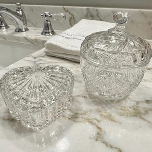 May include: Two clear glass decorative containers. One is heart-shaped with a lid, and the other is a round jar with a lid. Both have a cut glass design. They are on a marble countertop.