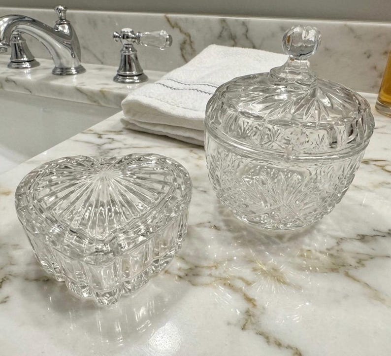 May include: Two clear glass containers with lids. One container is heart-shaped with vertical ribbed detailing. The second container is round with a faceted starburst pattern and a small knob handle on the lid. Both pieces are made of pressed glass.