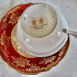 May include: A white teacup with a gold handle rests upside down on a saucer and a red and gold plate. The red plate has a gold floral design. The teacup and saucer are white with gold trim. The teacup is marked "Paragon" and "Fine Bone China England Regd."