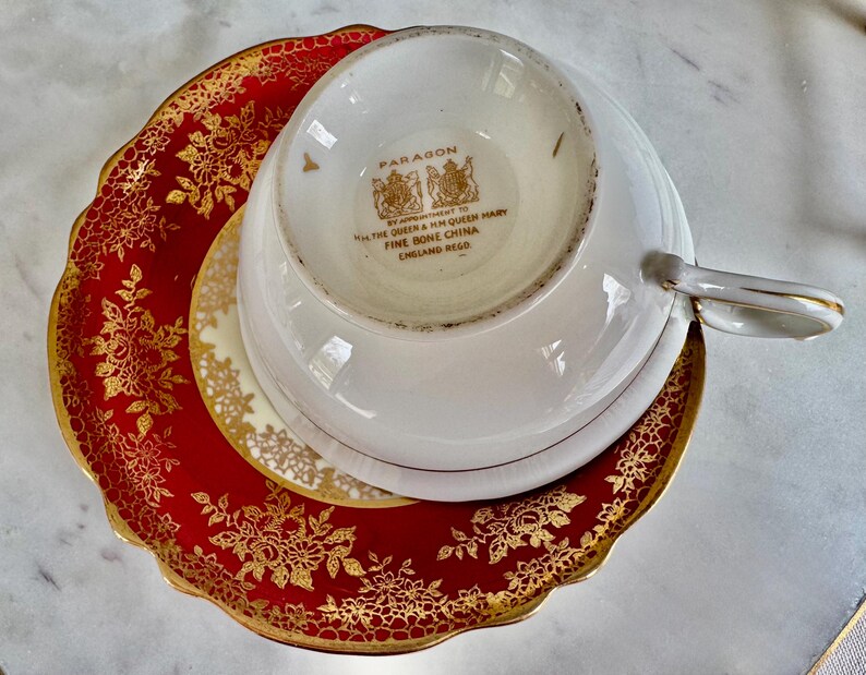 May include: A white teacup with a gold handle rests upside down on a saucer and a red and gold plate. The red plate has a gold floral design. The teacup and saucer are white with gold trim. The teacup is marked "Paragon" and "Fine Bone China England Regd."