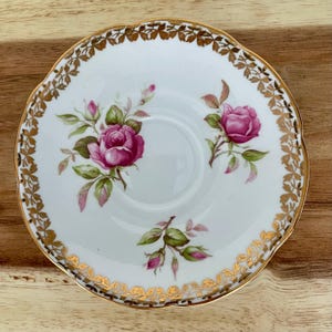 Puede incluir: Platillo de porcelana blanca con borde dorado festoneado y dise&ntilde;o floral de rosas. El platillo presenta rosas rosas con hojas verdes y detalles dorados. El dise&ntilde;o est&aacute; centrado en una superficie de madera.