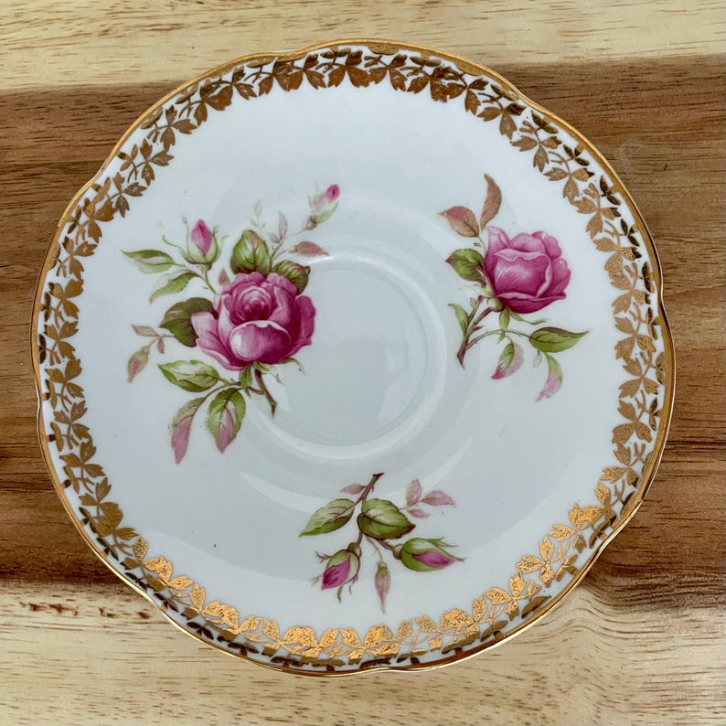 Puede incluir: Platillo de porcelana blanca con borde dorado festoneado y dise&ntilde;o floral de rosas. El platillo presenta rosas rosas con hojas verdes y detalles dorados. El dise&ntilde;o est&aacute; centrado en una superficie de madera.