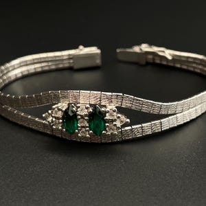 May include: A silver bracelet with a double row of linked squares. The bracelet has a central design featuring two emerald green stones surrounded by smaller clear stones.
