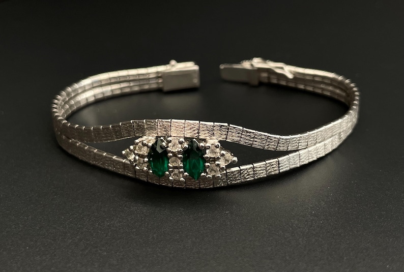 May include: A silver bracelet with a double row of linked squares. The bracelet has a central design featuring two emerald green stones surrounded by smaller clear stones.