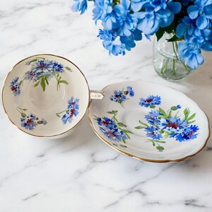 May include: A vintage teacup and saucer set with a floral pattern featuring blue and white cornflowers. The set has a gold trim around the edges.
