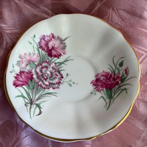 May include: A white porcelain saucer with a scalloped edge and a gold-trimmed rim. The center features a floral pattern of pink and white carnations with green stems and leaves arranged in two distinct clusters on the left and right sides.
