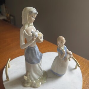 K&ouml;nnte beinhalten: Zwei Porzellanfiguren von Frauen auf einem herzf&ouml;rmigen Marmortablett. Eine Frau h&auml;lt einen Korb mit einem Vogel, die andere Blumen. Die Figuren sind in Wei&szlig;, Blau und Grau gekleidet.