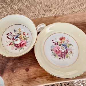 May include: A vintage teacup and saucer set with a floral design. The cup and saucer are both cream colored with a gold trim. The floral design features pink, red, yellow, and purple flowers.