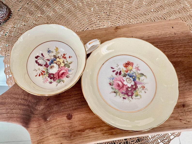 May include: A vintage teacup and saucer set with a floral design. The cup and saucer are both cream colored with a gold trim. The floral design features pink, red, yellow, and purple flowers.
