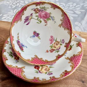May include: A vintage teacup and saucer set. The white porcelain cup and saucer feature a pink fish scale pattern and gold trim. The cup and saucer are decorated with floral bouquets in shades of pink, purple, and yellow.