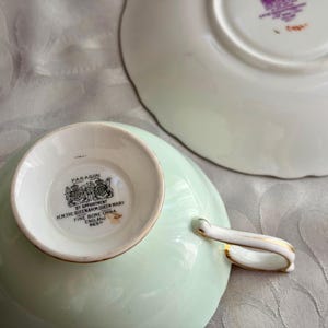 May include: A pale green bone china teacup and saucer set. The underside of the teacup features a black stamp reading Paragon, By Appointment H.M. The Queen & H.M. Queen Mary, Fine Bone China, England. The saucer has a matching stamp in purple.