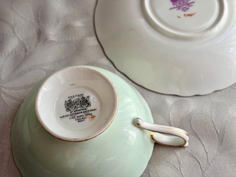May include: A pale green bone china teacup and saucer set. The underside of the teacup features a black stamp reading Paragon, By Appointment H.M. The Queen & H.M. Queen Mary, Fine Bone China, England. The saucer has a matching stamp in purple.