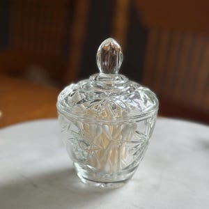May include: A clear glass lidded jar featuring a faceted starburst pattern on the body and lid. The jar has a rounded base and a pointed finial handle on top. Inside the container, a collection of white cotton swabs is clearly visible.